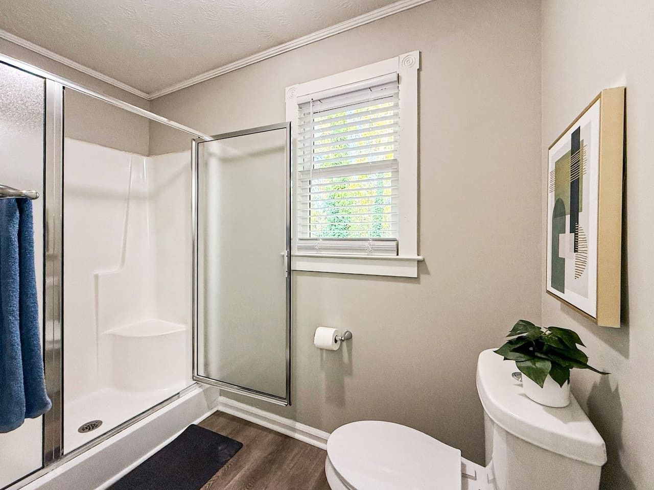 Un baño con ducha de vidrio, inodoro, ventana con persianas y un cuadro en la pared.