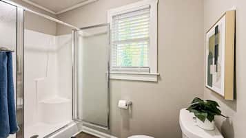 Un baño con ducha de vidrio, inodoro, ventana con persianas y un cuadro en la pared.