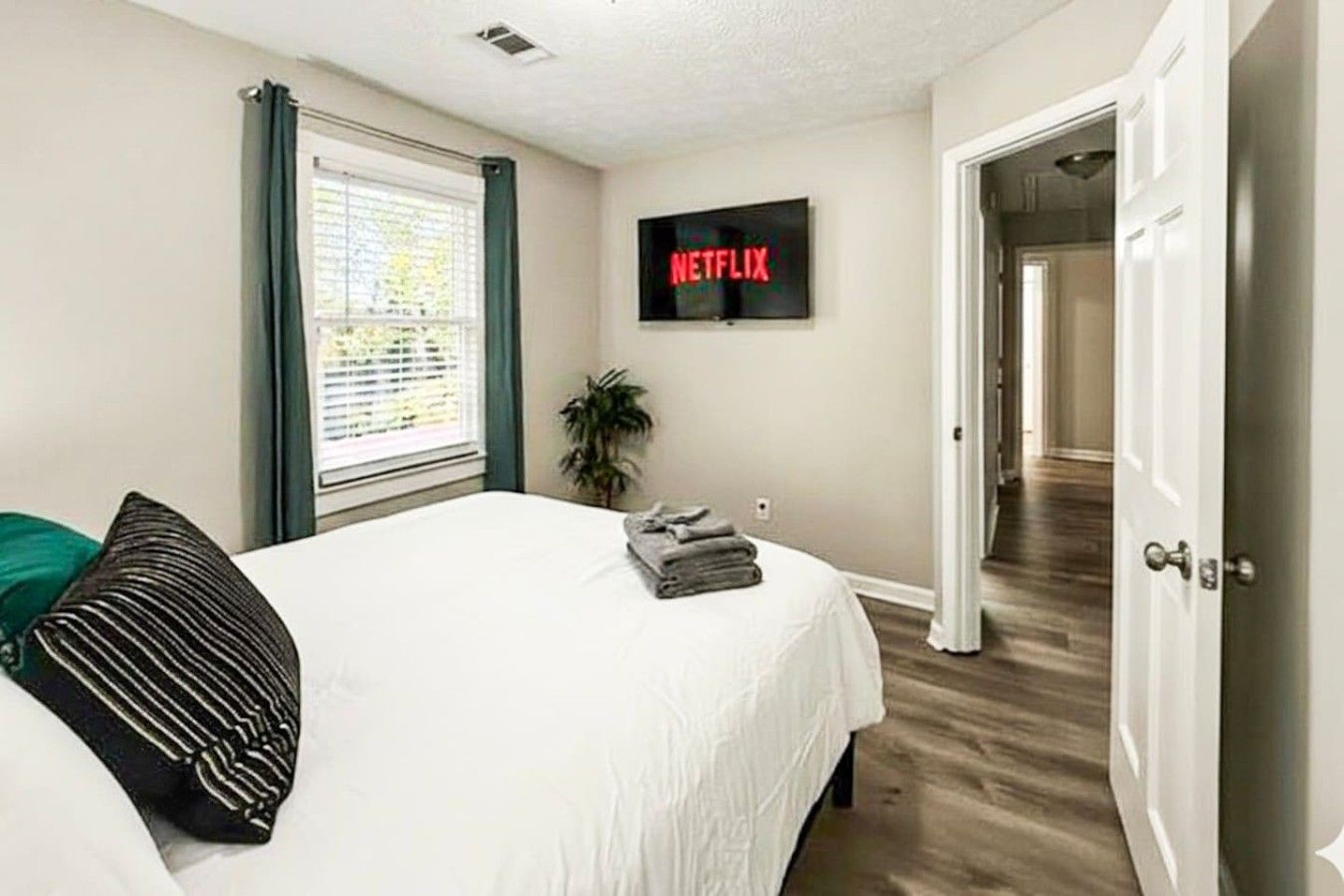 A bedroom with a bed, a television displaying Netflix, a window with blinds, and a door leading to another room.