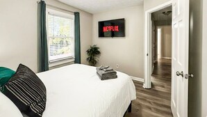 Een slaapkamer met een bed, een televisie waarop Netflix wordt afgespeeld, een raam met jaloezieën en een deur naar een andere kamer.