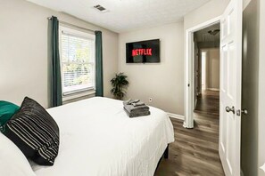 Une chambre à coucher avec un lit, un téléviseur diffusant Netflix, une fenêtre avec des stores et une porte donnant sur une autre pièce.
