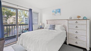Une chambre à coucher avec un lit, une commode, une fenêtre donnant sur un balcon et un tableau accroché au mur.