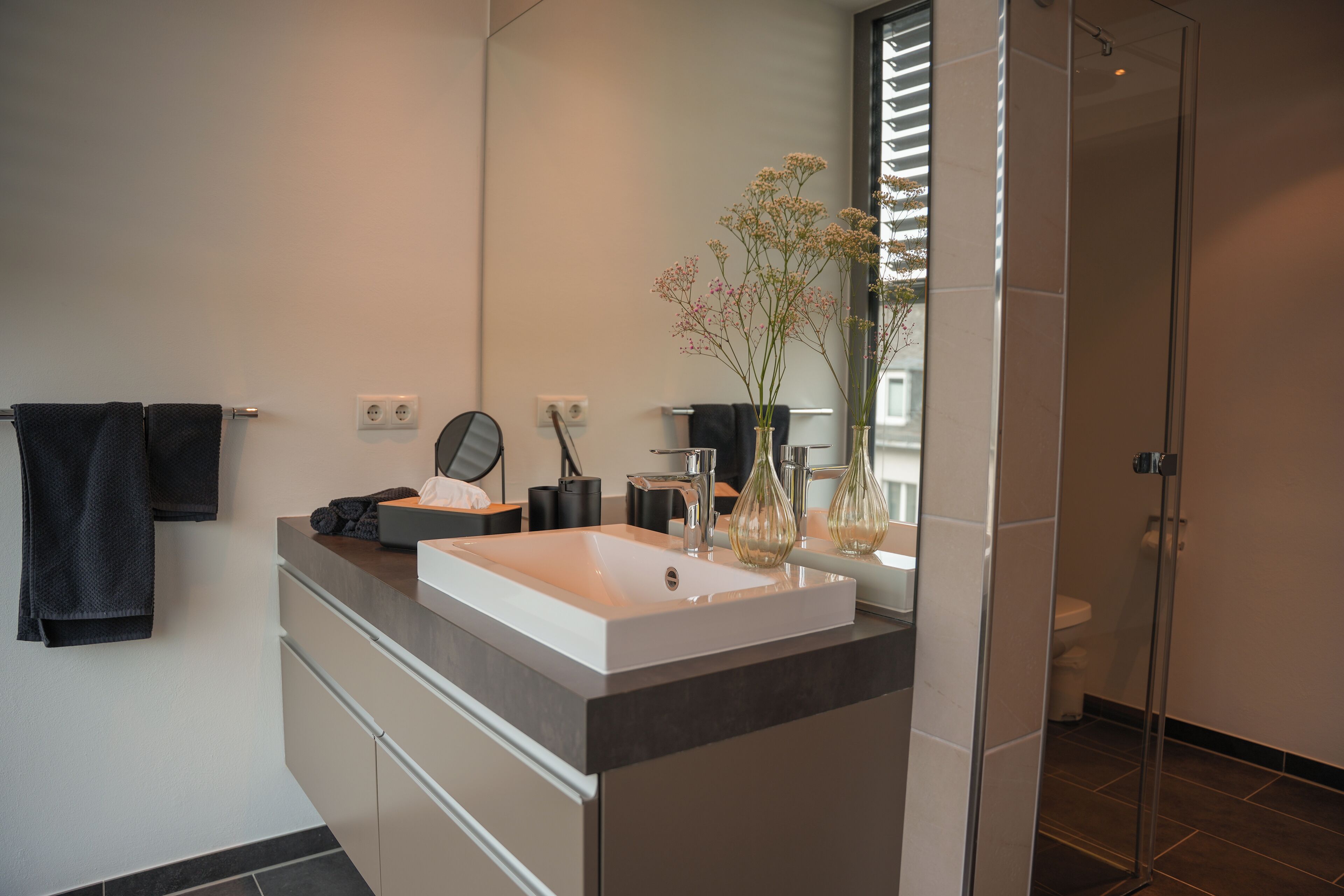 Une salle de bain moderne avec un grand miroir, un lavabo blanc et un vase rempli de fleurs.