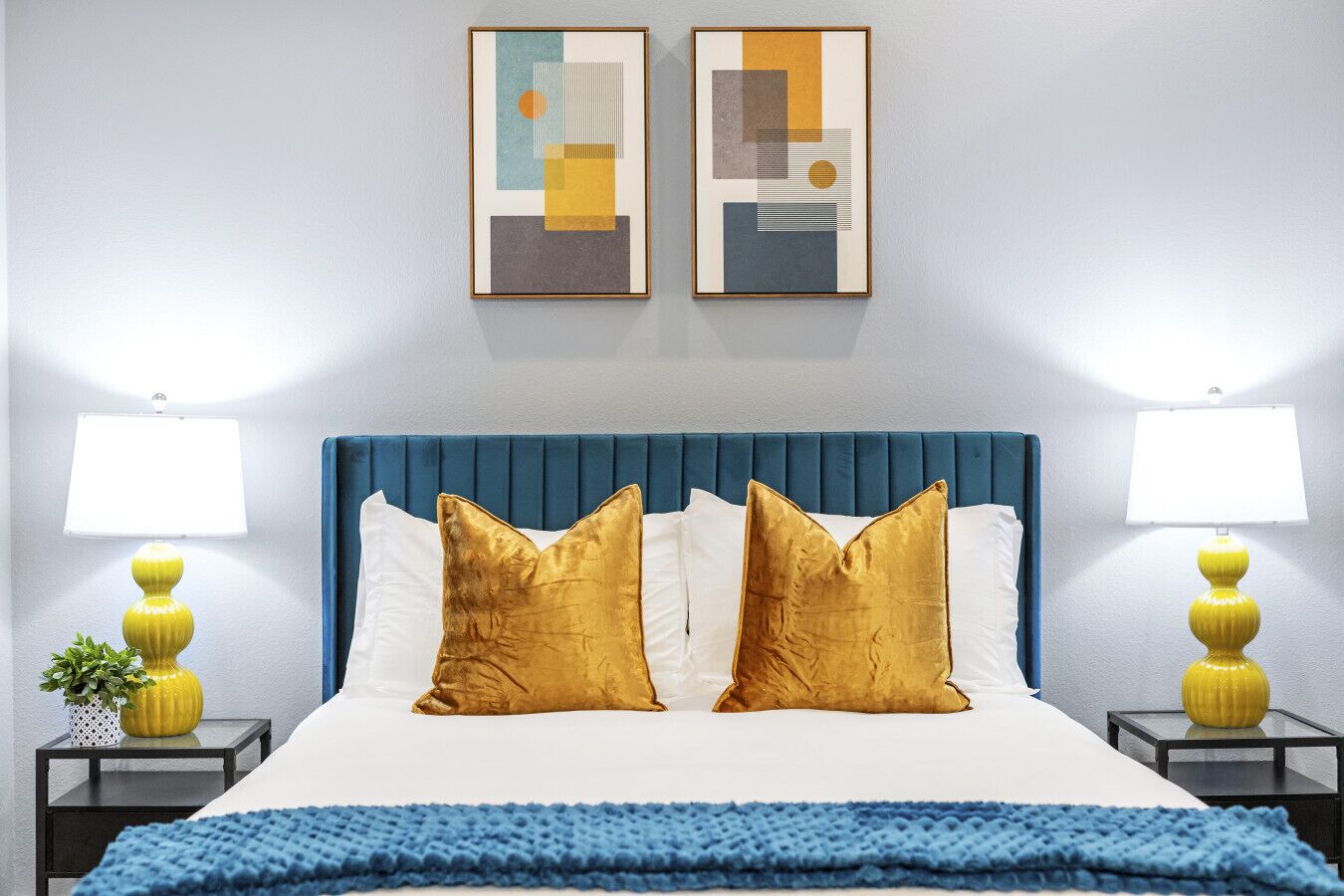 A hotel room with a blue upholstered headboard, two gold pillows, white bedding, and two yellow lamps on side tables.