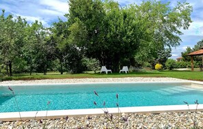 Une piscine entourée d’un bord de galets, avec deux chaises longues blanches placées sous un grand arbre.