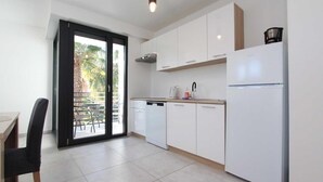 Cozinha moderna com armários brancos, lava-louças, pia e geladeira. Há uma mesa de jantar com cadeiras e vista para palmeiras através de uma porta de vidro.