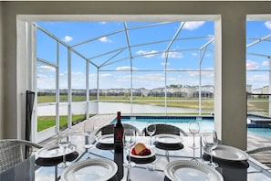 A glass-enclosed patio with a pool view, dining table set with plates and wine glasses, and a bottle of wine. - Family 9BR Lakeview Home w/ Pool, Jacuzzi & Game Room on ChampionsGate Resort! (Kissimmee)
