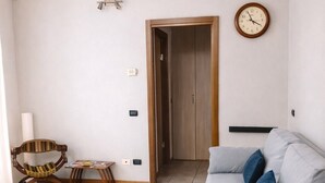 A living room with a grey sofa, a wooden door, a wall clock, and a small table with a chair. - La Corte Guesthouse – Independent stay with private garden near Milan (Cornate d'Adda)