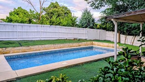 Une piscine rectangulaire bordée de pierres, entourée de gazon synthétique et d’une clôture blanche.