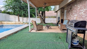 Un jardin avec une piscine, du gazon synthétique, des meubles en osier et un barbecue.