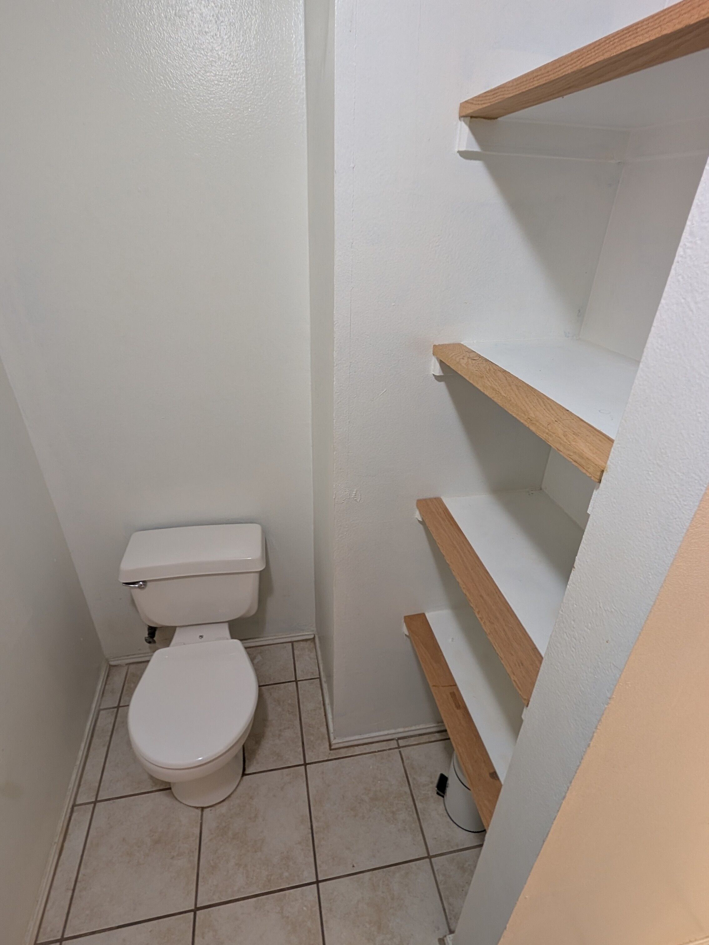 Ein kleines Badezimmer mit Toilette, Fliesenboden und Holzregalen.