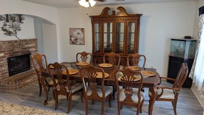 Une salle à manger avec une cheminée en briques, des chaises en bois, une table à manger avec un plateau en verre et un buffet en bois.