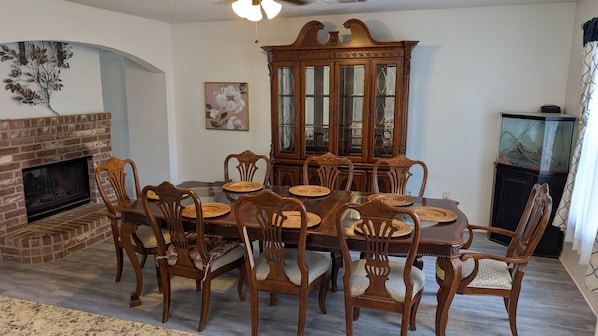Uma sala de jantar com lareira de tijolos, cadeiras de madeira, mesa de jantar com tampo de vidro e um armário de madeira.