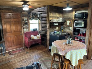 A rustic cabin interior with a wooden door, a red bench, a dining table with a checkered tablecloth, a kitchenette with a microwave and toaster, and a window with curtains.