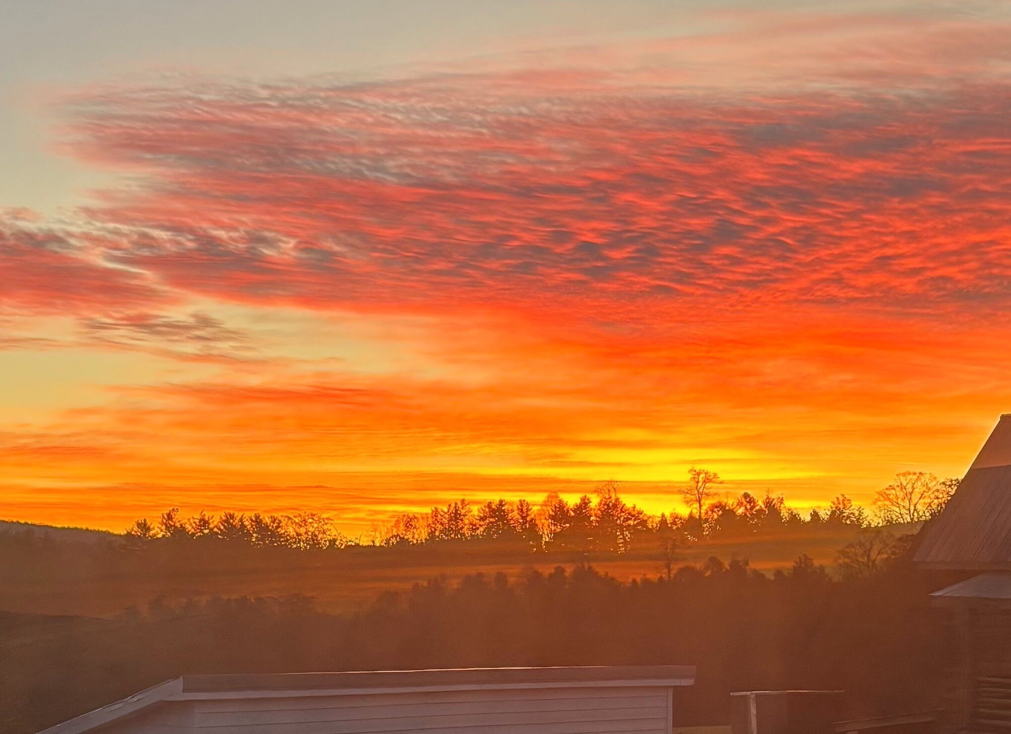 Auringonlasku maaseutumaisemassa, jossa näkyy puiden ja talon siluetit etualalla.