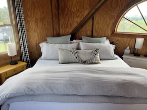 A bedroom with a bed, pillows, a nightstand, a lamp, and a window with a view of the outdoors.