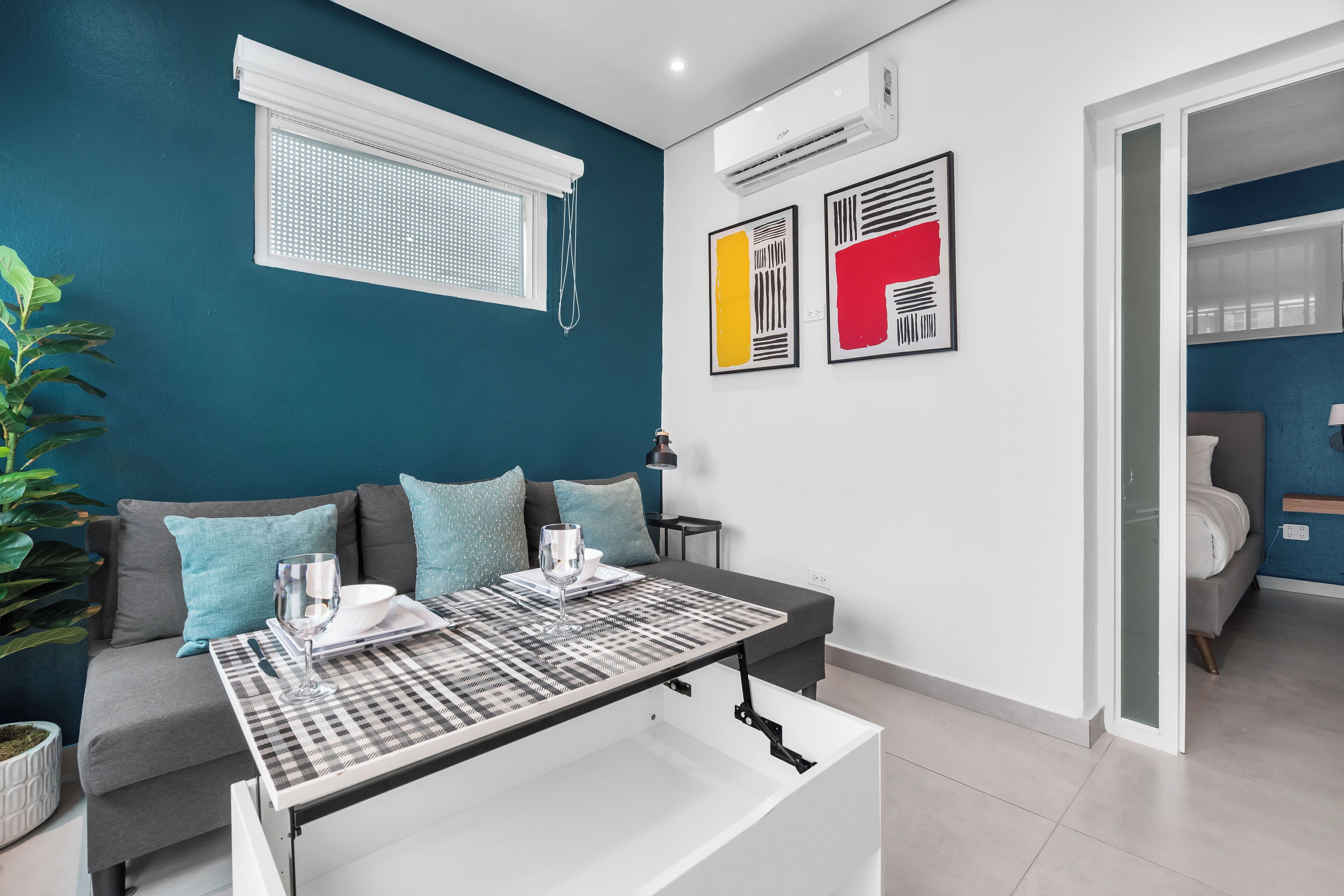 Un salon moderne avec un canapé, une table basse et une table à manger. Il y a une fenêtre avec des stores, une plante et des œuvres d’art accrochées au mur.
