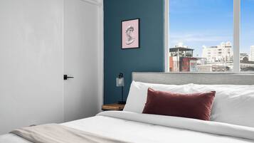 Cama bem arrumada com almofadas brancas e vermelhas, mesa de cabeceira com abajur e quadro emoldurado numa parede azul.