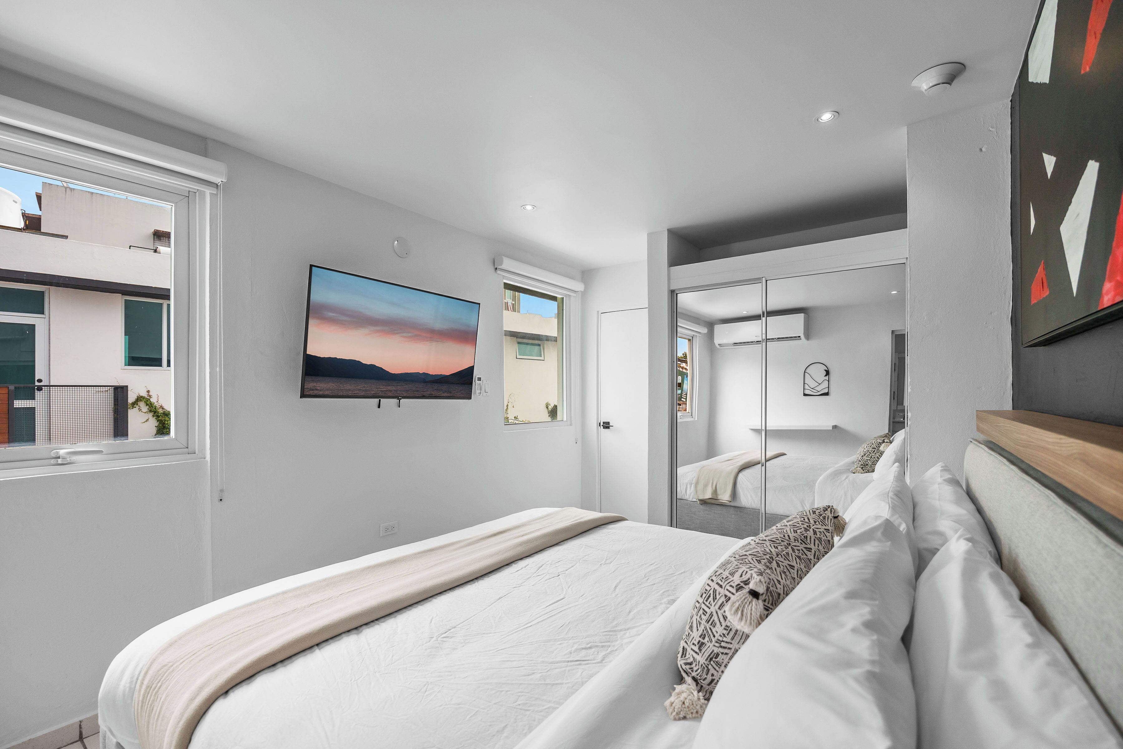 A modern bedroom with a large bed, a TV mounted on the wall, and a view of the outside through a window.