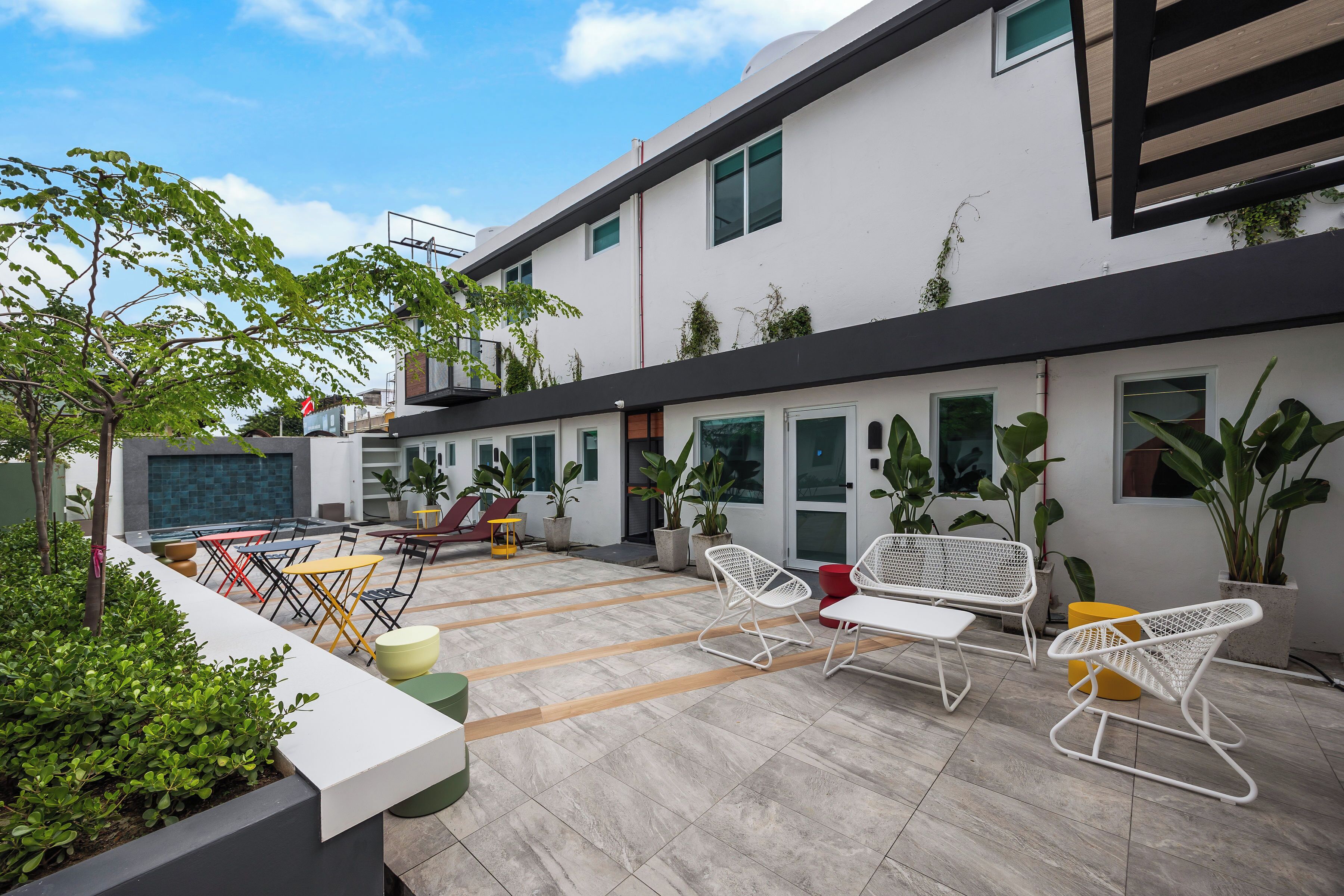Un patio moderno al aire libre con mobiliario, plantas en macetas y un edificio con ventanas amplias.