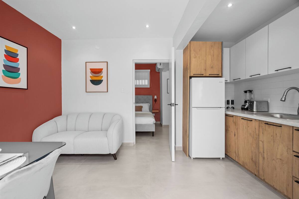 Una cucina moderna con mobili bianchi, un frigorifero e un lavello. Nell'ambiente adiacente, si trova un divano bianco e un letto.