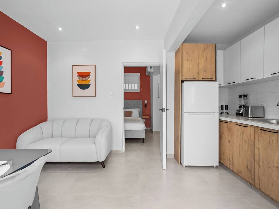 Eine moderne Küche mit weißen Schränken, einem Kühlschrank und einem Spülbecken. Im angrenzenden Raum befinden sich eine weiße Couch und ein Bett.