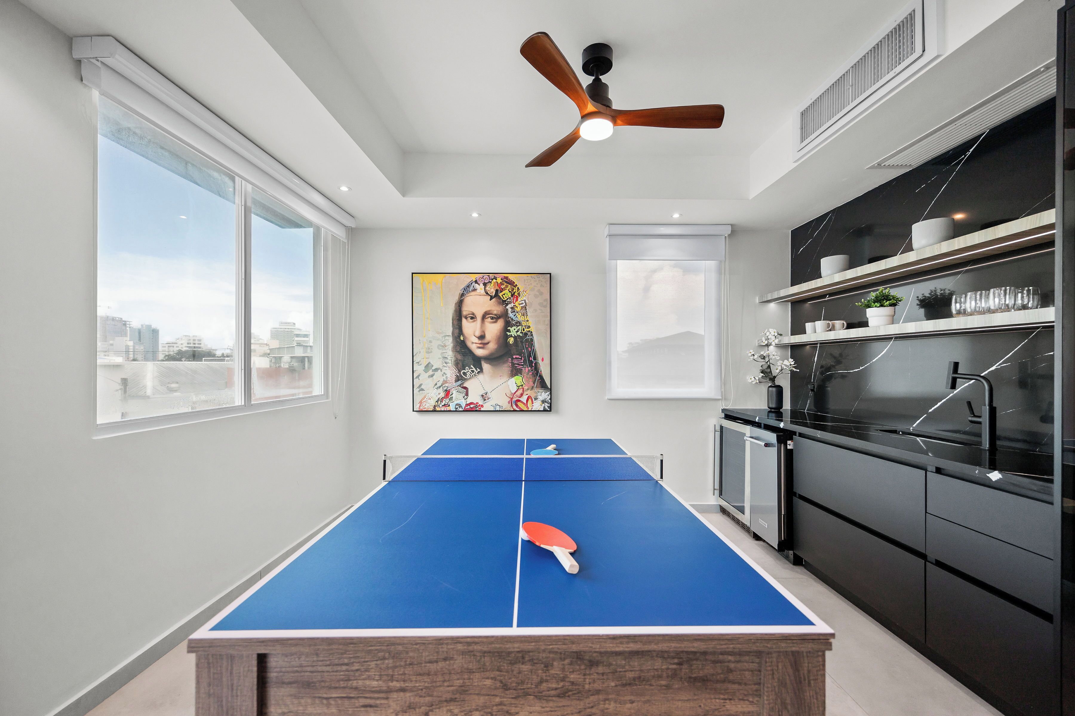 Une table de ping-pong dans une cuisine moderne avec vue sur la ville, un ventilateur de plafond et un tableau accroché au mur.