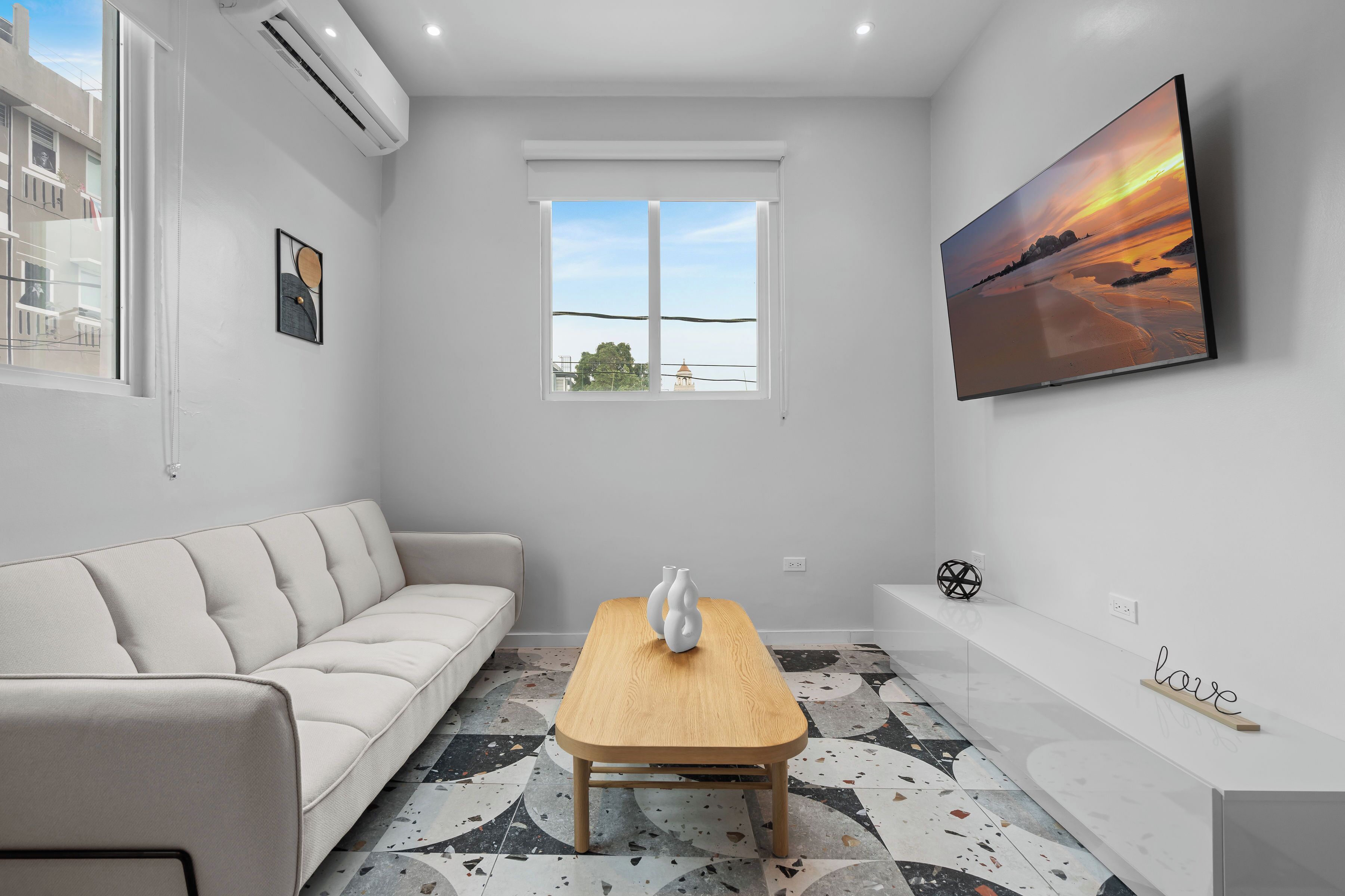 A modern living room with a sofa, a wooden coffee table, a TV mounted on the wall, and a window with a view of the outside.