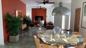 Un comedor moderno con mesa de vidrio y sillas de madera, ventilador de techo y una planta grande.