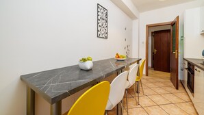 Uma sala de jantar com uma mesa de mármore, cadeiras brancas e amarelas, uma tigela de frutas e um objeto decorativo na parede.