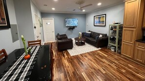 A living room with a dark brown leather sofa, a wooden dining table with a black and white checkered tablecloth, and a television mounted on the wall.