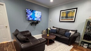 A living room with a dark brown leather sofa, a smaller armchair, a coffee table, a TV mounted on the wall, and a framed painting.