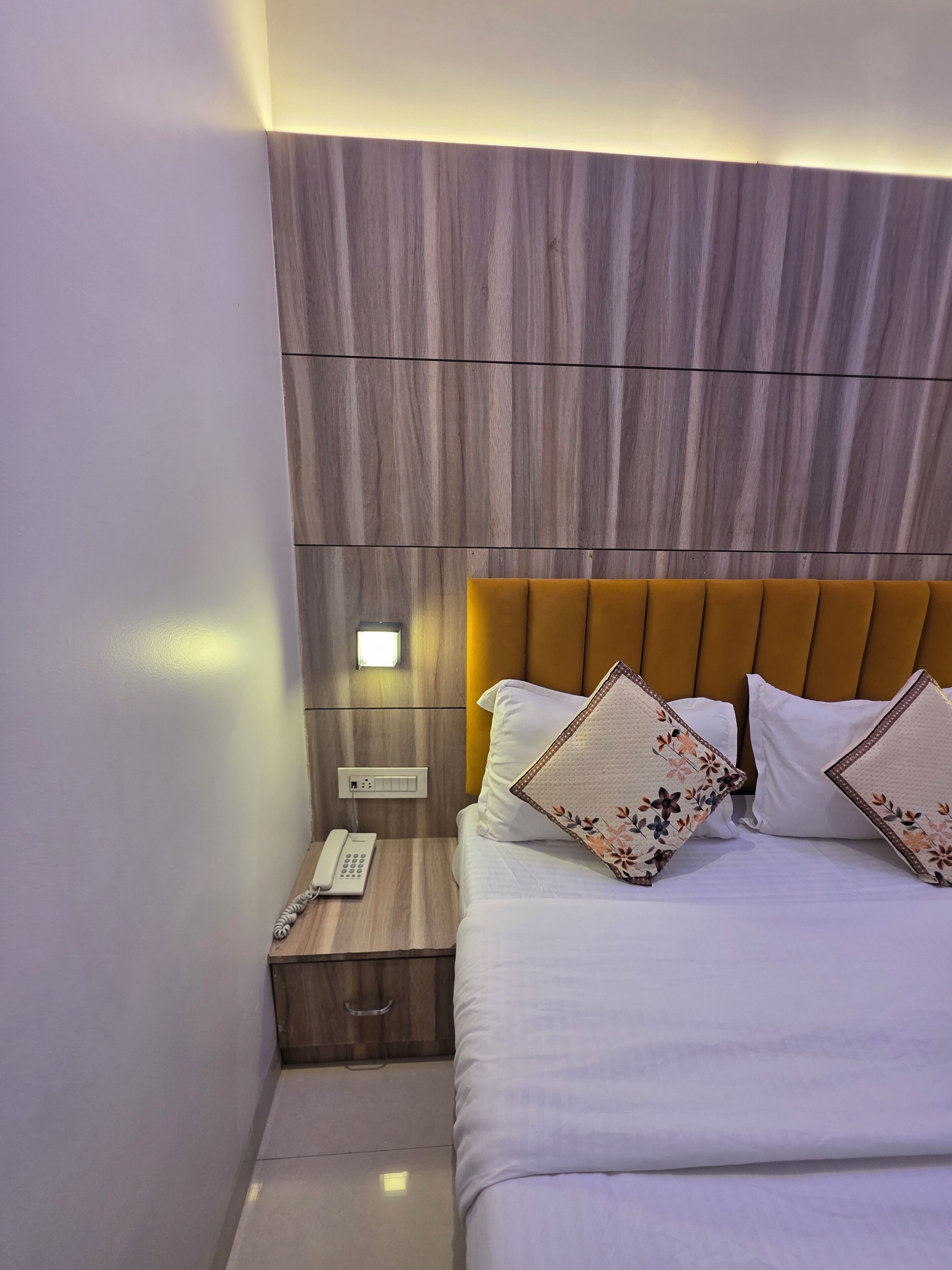 Una cama bien hecha con sábanas blancas y almohadas estampadas con motivos florales, un cabecero de madera y un teléfono en la mesita de noche.