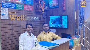 A hotel reception desk with a wooden finish, a digital screen displaying information, and a sign indicating check-in and check-out times. - Hotel Wellingten (Mumbai)