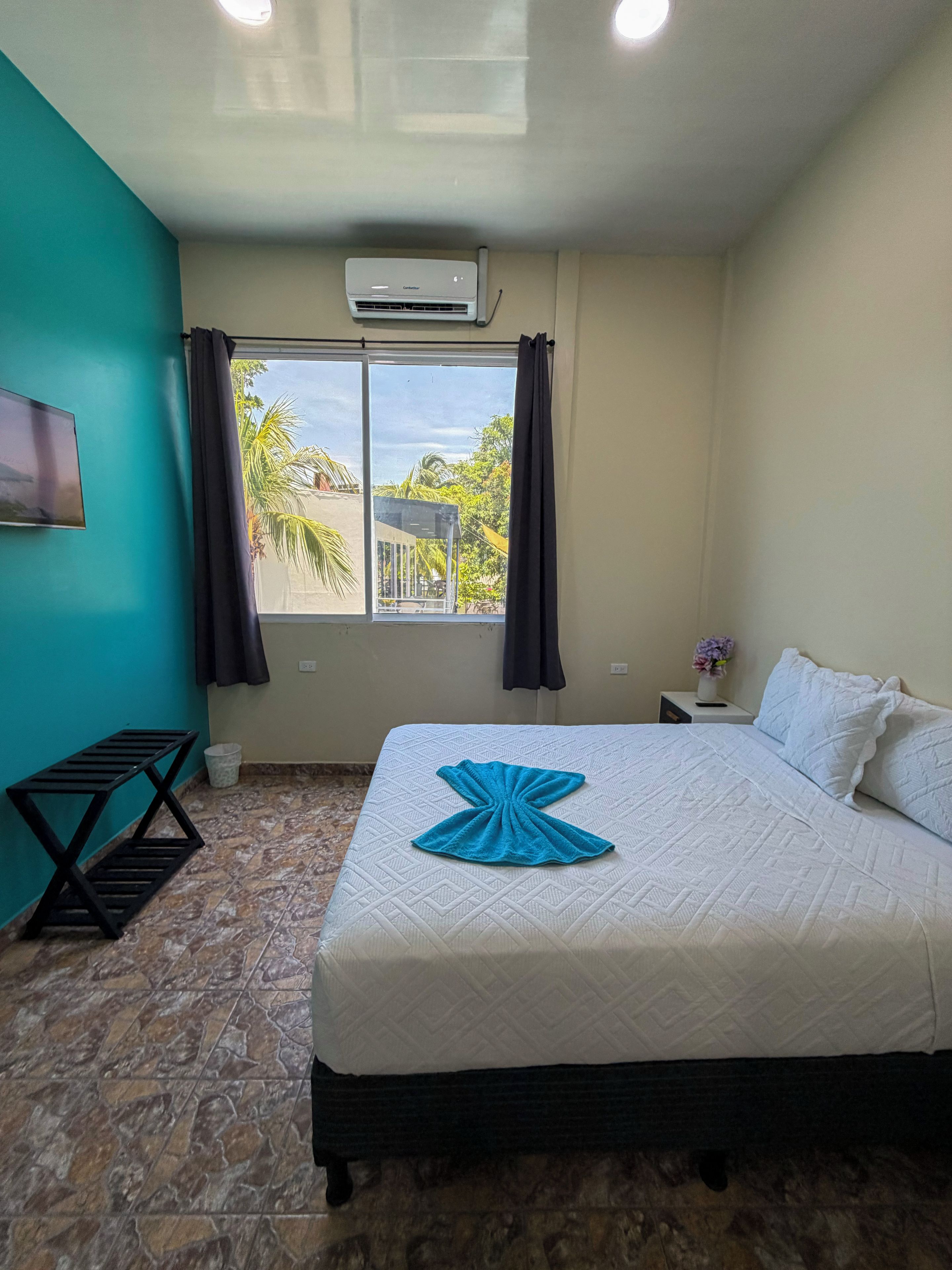 A hotel room with a bed, a small table, a wall-mounted TV, a window with curtains, and an air conditioning unit.