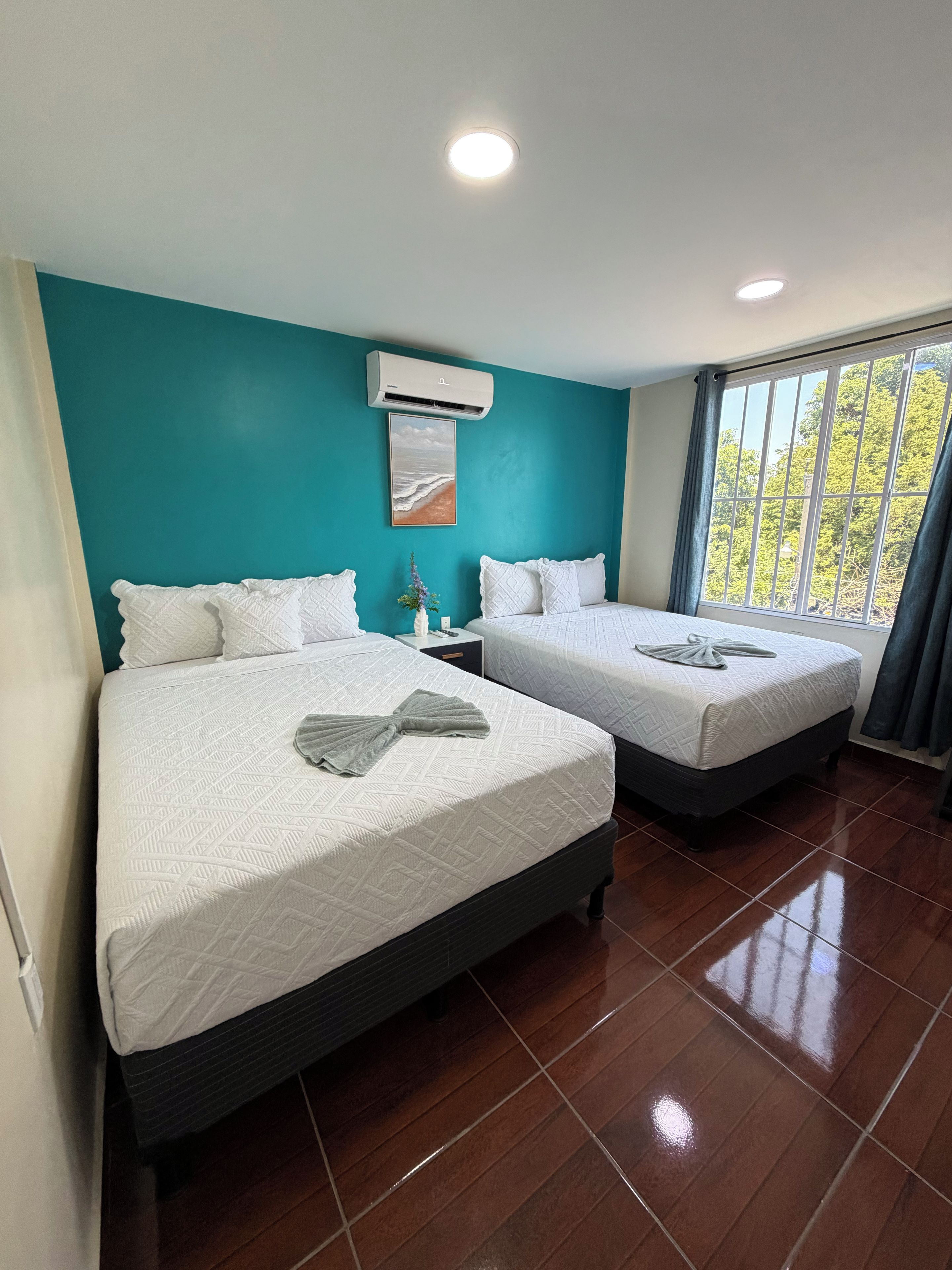 A hotel room with two beds, an air conditioning unit, a painting, and a window with curtains.