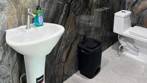 A bathroom with a white sink, a black trash bin, a toilet, and a dark blue shelf unit against a marble wall. - Mi Tierra Surf City Hotel and Restaurant (La Libertad)