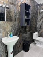 Un baño con un lavamanos blanco, un cubo de basura negro, un inodoro y un estante azul oscuro sobre una pared de mármol.