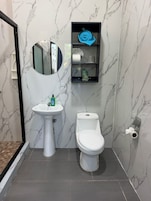 Un baño con lavamanos pedestal blanco, inodoro, espejo y repisa con artículos de tocador.