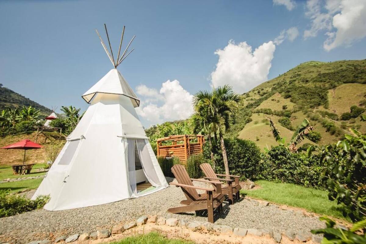 A white teepee with a door, surrounded by a gravel area and wooden chairs, set against a hilly landscape with palm trees and a clear sky.