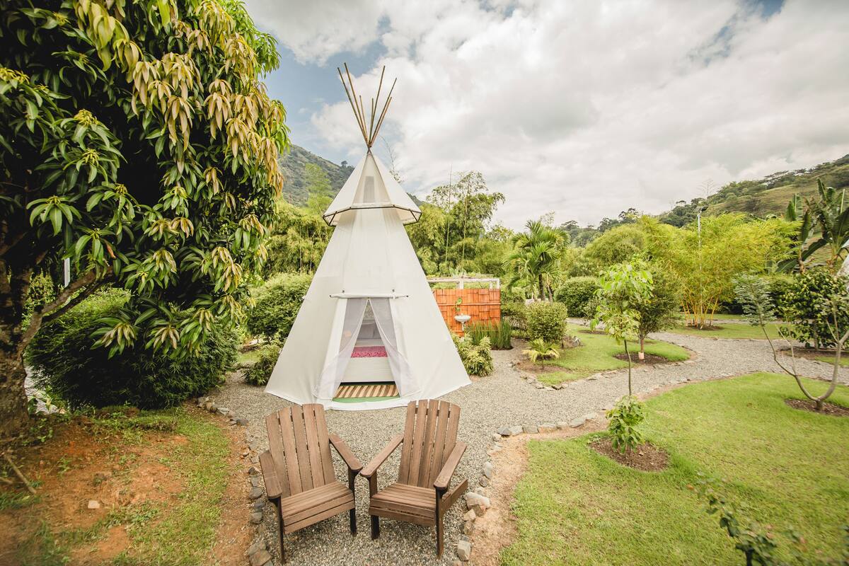 A white teepee tent with two wooden chairs in front, surrounded by lush greenery and a gravel path.