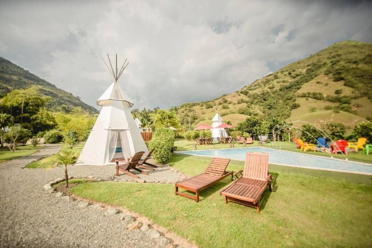 A white teepee, a pool, and lounge chairs in a grassy area with hills in the background.