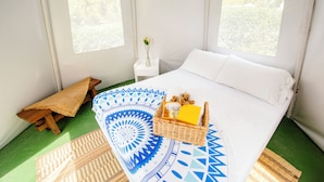 A neatly arranged bedroom with a bed, a small wooden bench, a wicker basket with toiletries, and a window with a view of greenery. - Cerro Tusa Glamping  (Titiribí)