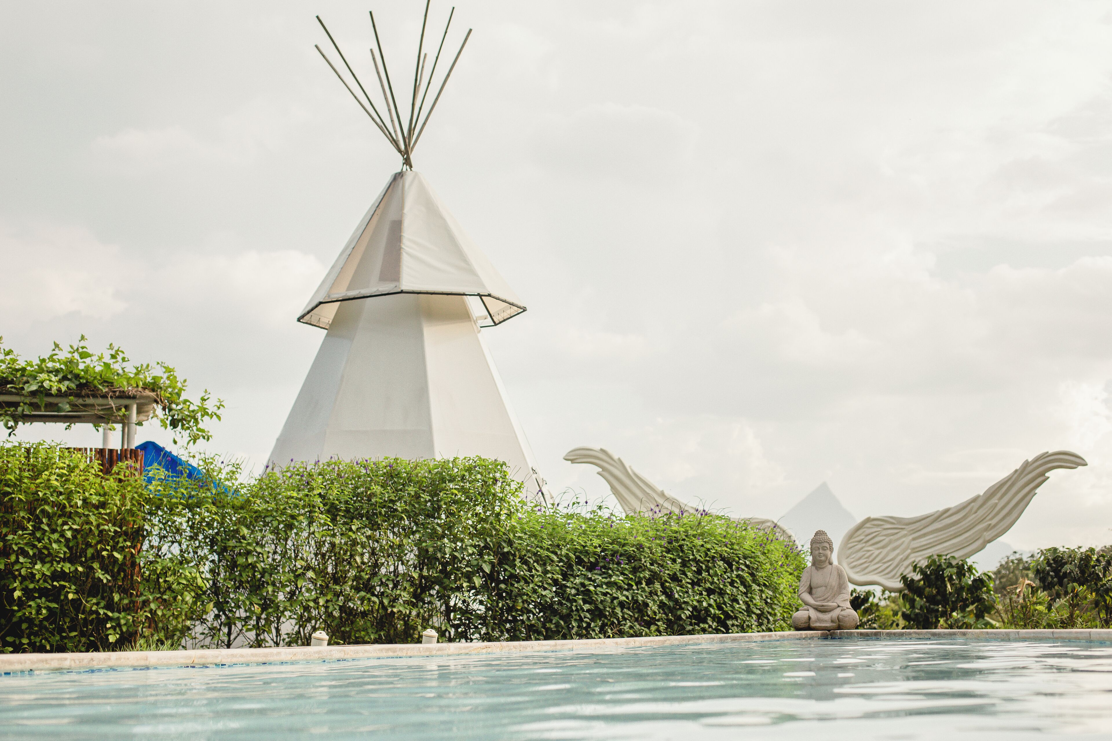 A swimming pool with a statue of a figure with wings, a white conical structure, and lush greenery.