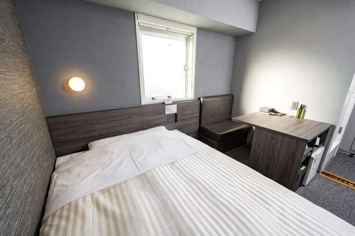 A hotel room with a bed, a desk, a chair, a window, and a wall-mounted light.
