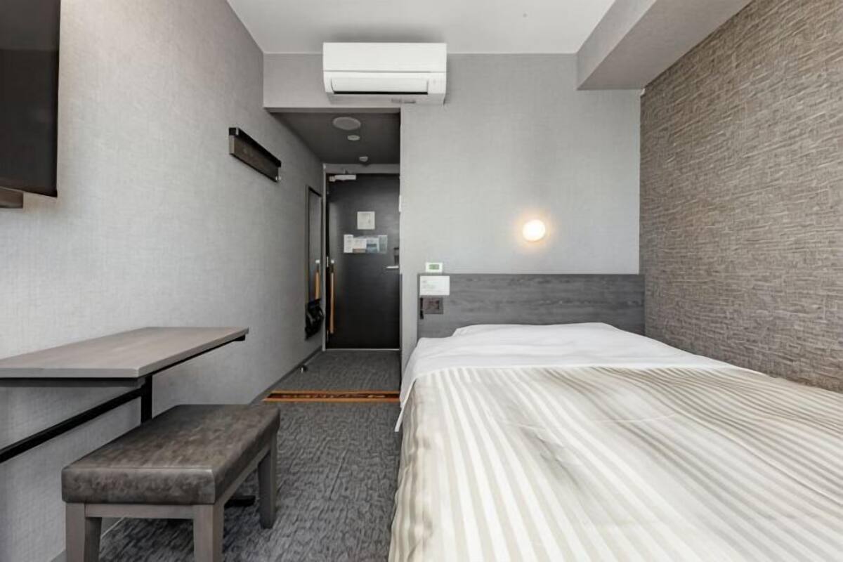 A hotel room with a bed, a desk, a bench, a wall-mounted air conditioner, and a door leading to a bathroom.