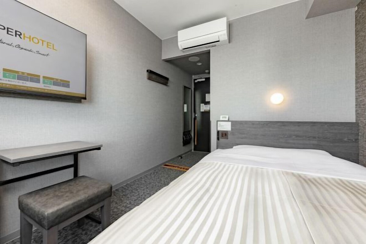 A hotel room with a bed, a desk, a bench, and a wall-mounted air conditioner.