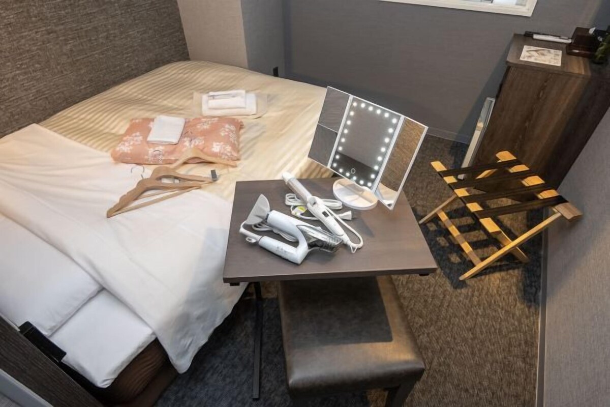 A neatly made bed with a white comforter and pillows, a small table with a mirror and vanity items, and a wooden chair.