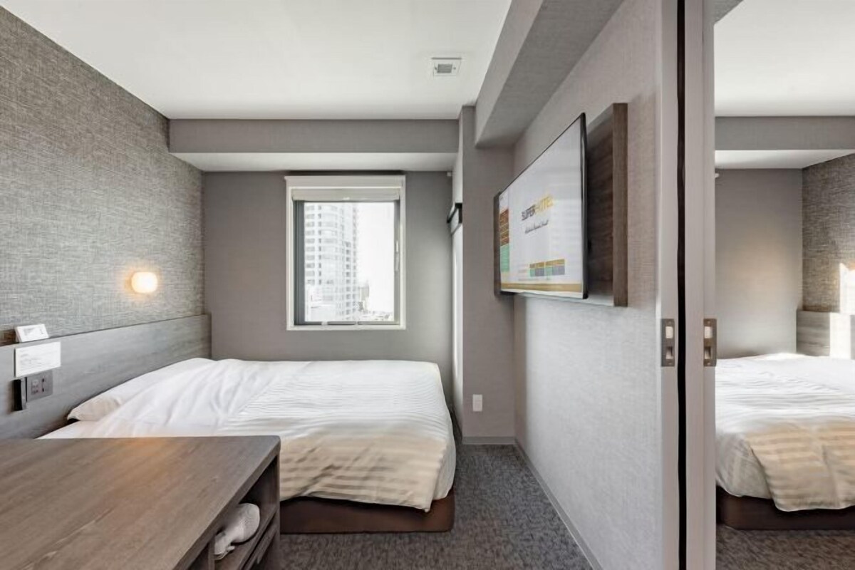 A hotel room with two beds, a desk, a window, and a view of a building.