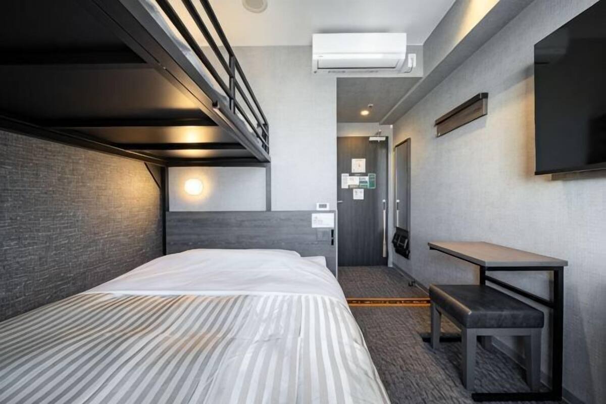 A hotel room with a bunk bed, a desk, a chair, a flat-screen TV, and a wall-mounted air conditioner.